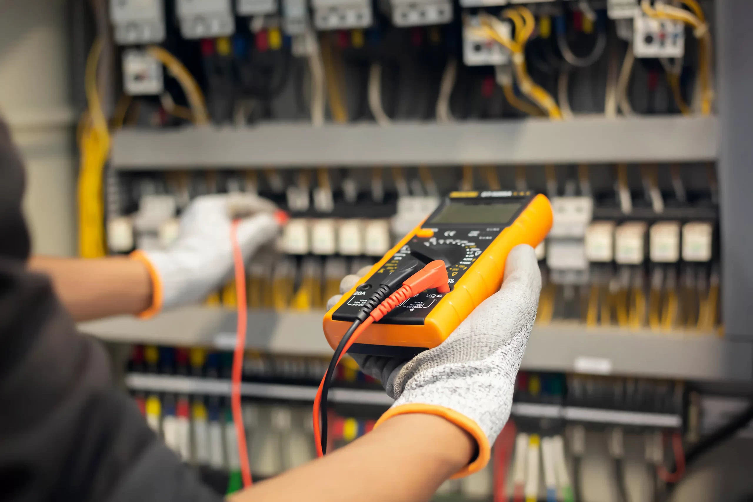 A person wearing white gloves holds an orange digital multimeter to perform crucial electrical safety checks on an electrical panel in a control room, surrounded by various wires and switches.