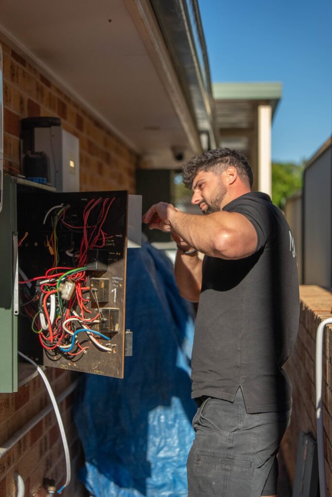 Electrician working on an old open switchboard with original wiring on the brick wall of a Cecil Hills home before the upgrade
