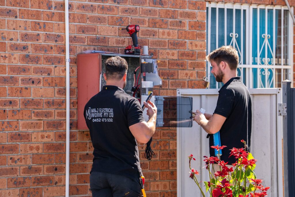 Two electricians working at an open switchboard enclosure on the brick wall of a Hoxton Park house